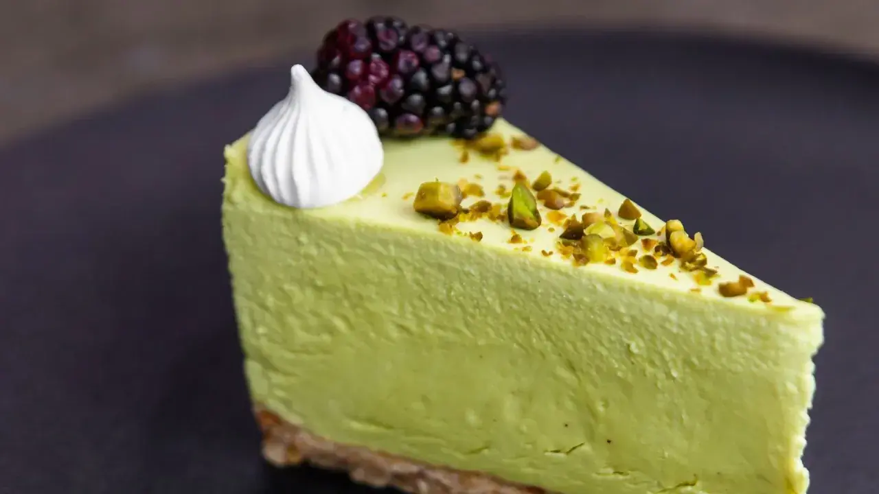 Pyszny sernik pistacjowy na zimno, udekorowany pianką, jeżyną i posypany pistacjami.