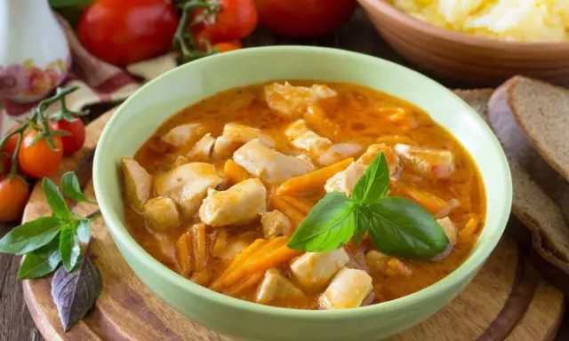 Aksamitny gulasz z piersią z kurczaka z marchewką, udekorowany bazylią. Obok kromki chleba i miska purée ziemniaczanego.