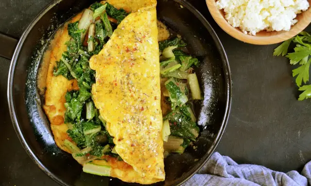 Omlet z serka wiejskiego z zielonymi warzywami na patelni. Obok miseczka z serkiem i natka pietruszki.