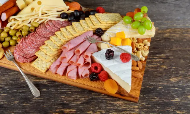 Apetyczna deska serów i wędlin: salami, szynka, ser żółty, pleśniowy, oliwki, winogrona, maliny, orzechy i krakersy.