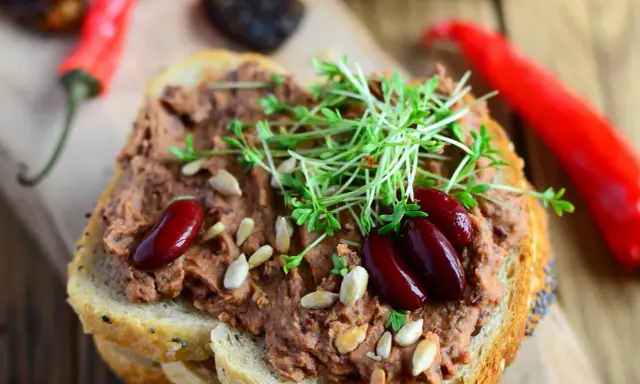 Pyszna pasta z czerwonej fasoli na chlebie, ozdobiona kiełkami i nasionami. Obok leżą czerwone papryczki chili.