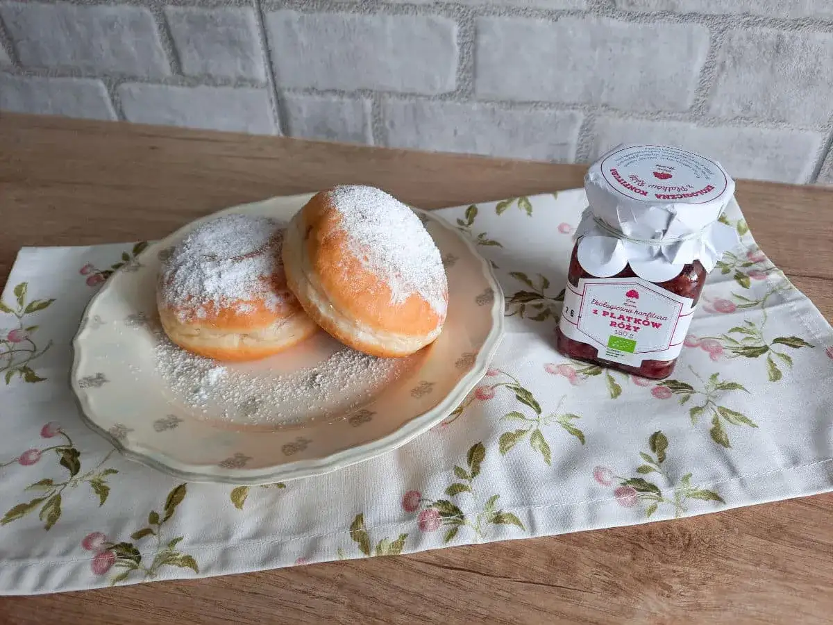 Pączki z przepisu Ewy Wachowicz, opr&oacute;szone cukrem pudrem, obok słoiczka konfitury z płatk&oacute;w r&oacute;ży.
